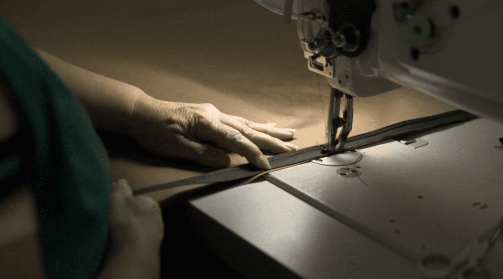 A close up of a persons hand guiding fabric through an industrial sewing machine stitching a zipper onto the edge of the material
